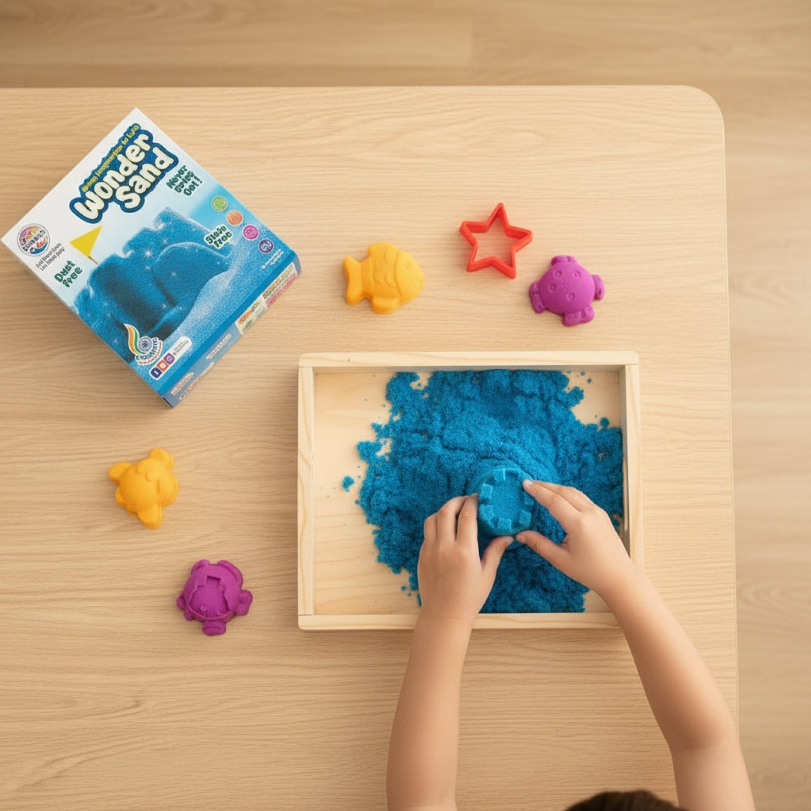 Children playing with blue kinetic sand in a wooden box on a wooden table, with a box of Wonder Sand visible.
